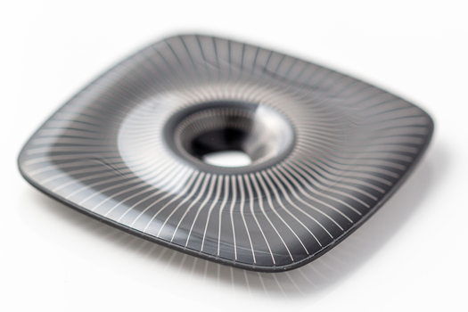 schwarzes quadratisches Bauteil mit radialen Linien und zentraler Öffnung